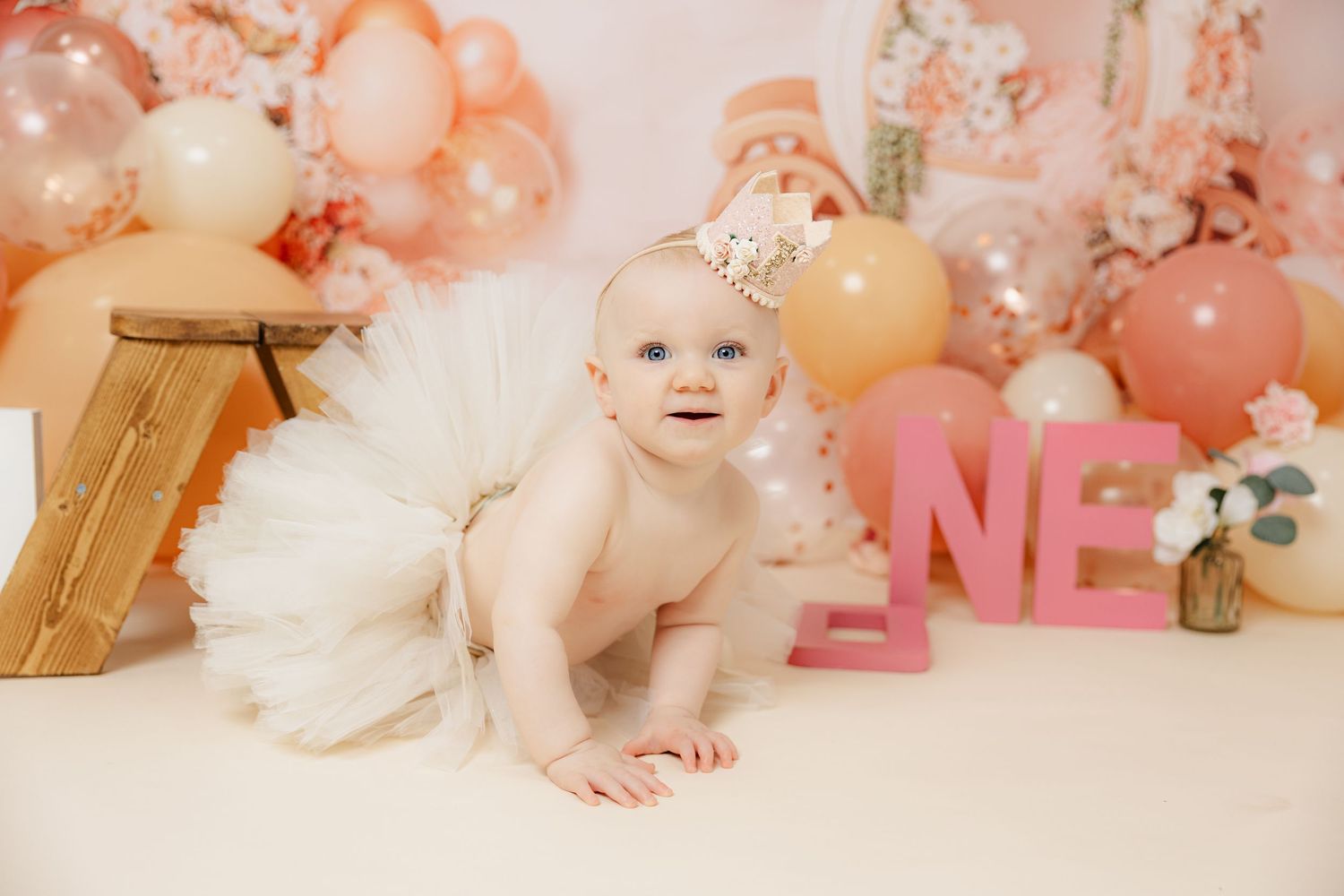 A baby girl in a white tutu crawls among pink balloons and ONE letters at a first birthday cake smash photoshoot.