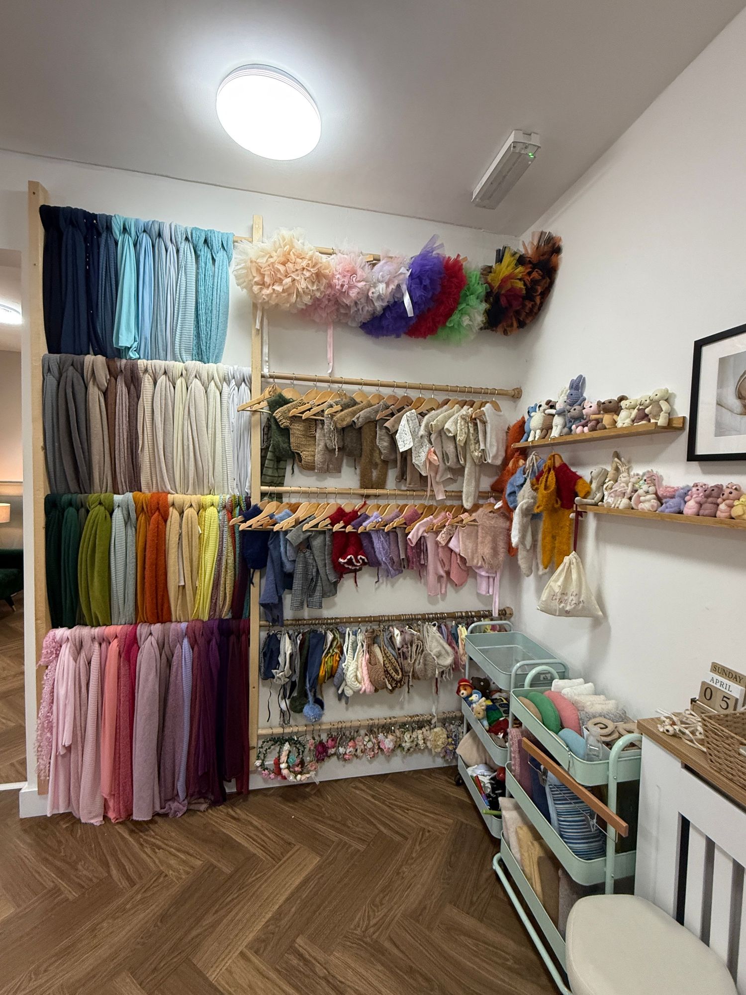 Colorful fabric swatches and baby clothing organized on wooden shelves in a bright photography studio prop room.