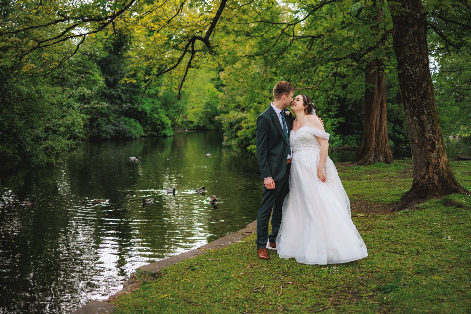 A bride and groom share a romantic kiss by a serene pond surrounded by lush green trees on their wedding day.