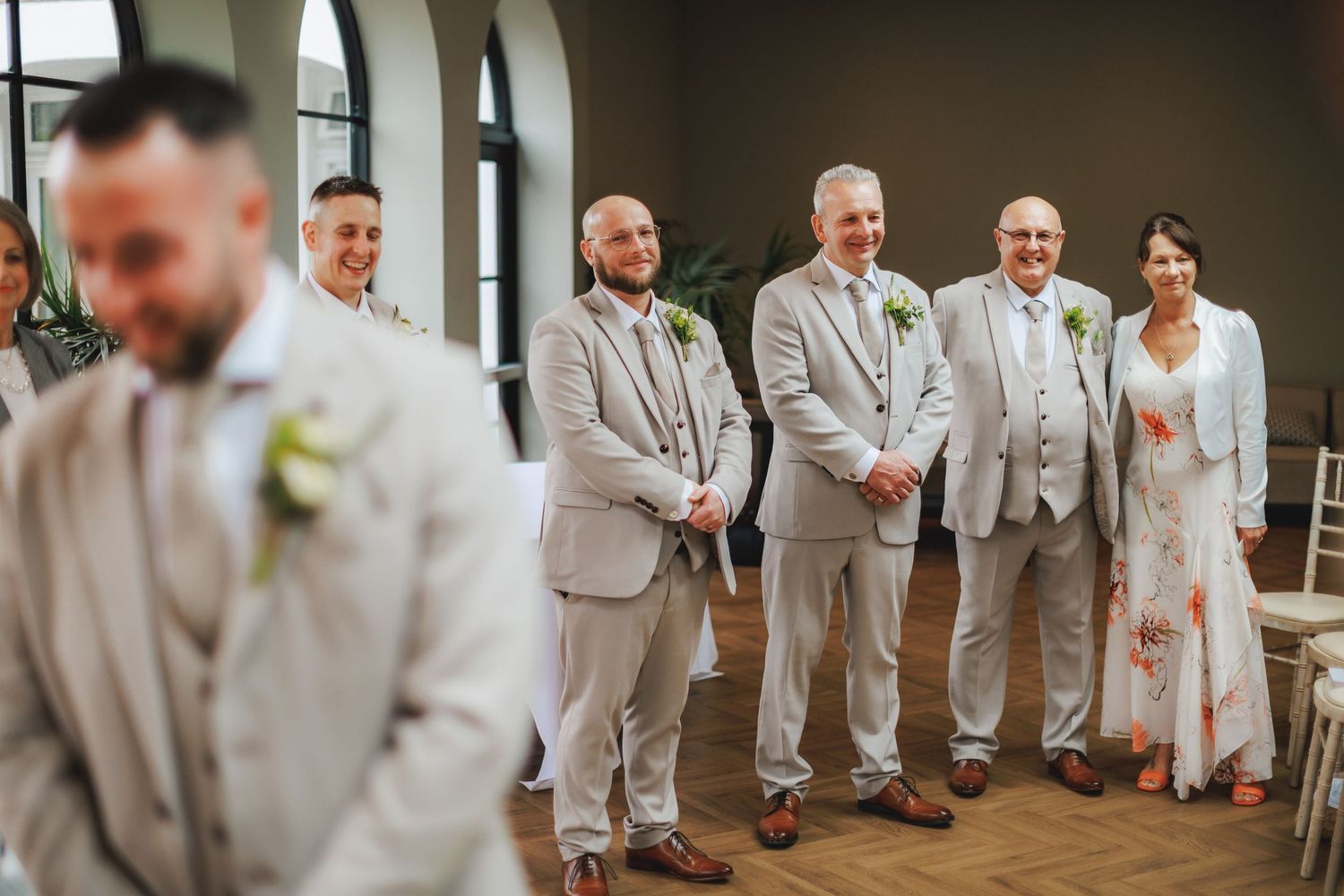 Groomsmen in light grey suits stand smiling at a wedding ceremony indoors, with a woman in a floral dress beside them.