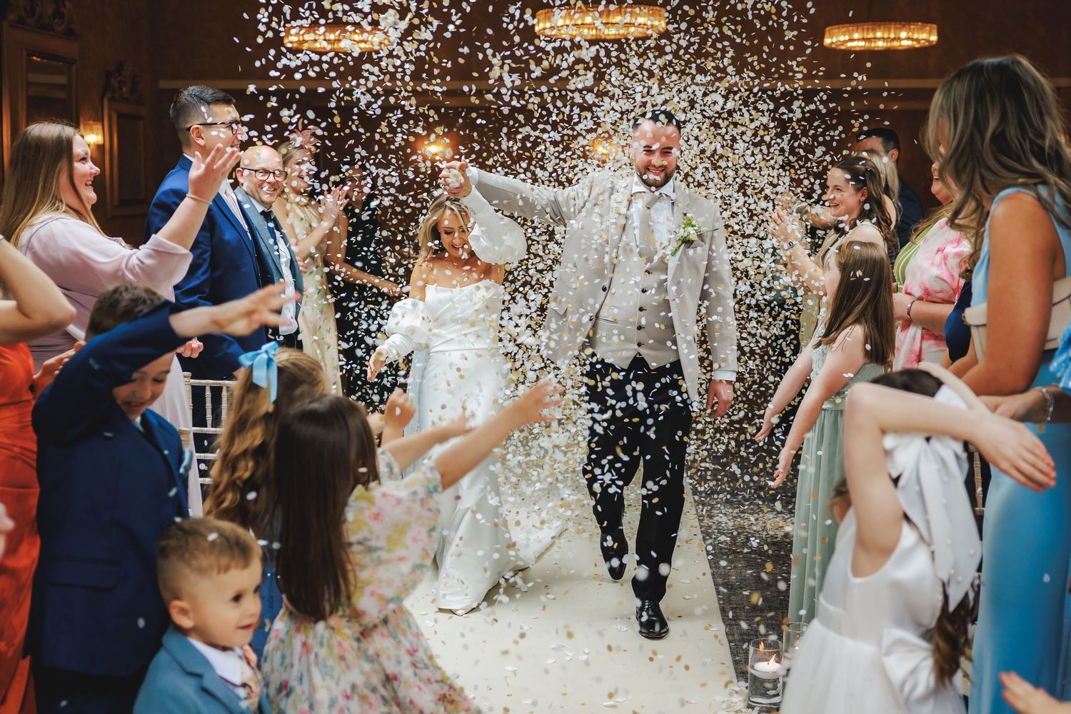 Bride and groom walk through confetti shower surrounded by cheering wedding guests in elegant venue.