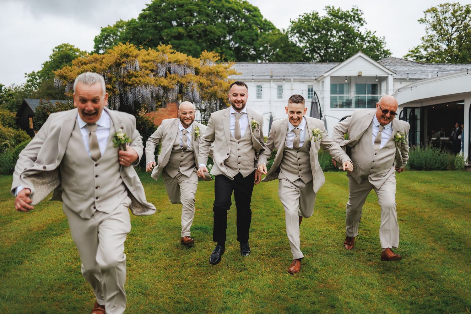 Groomsmen in light gray suits laughing and running together on green lawn outside elegant wedding venue.