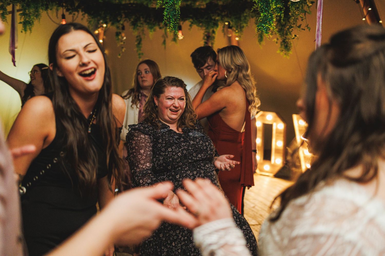 Women laughing and dancing joyfully at an indoor party with warm string lights and lush greenery decor.
