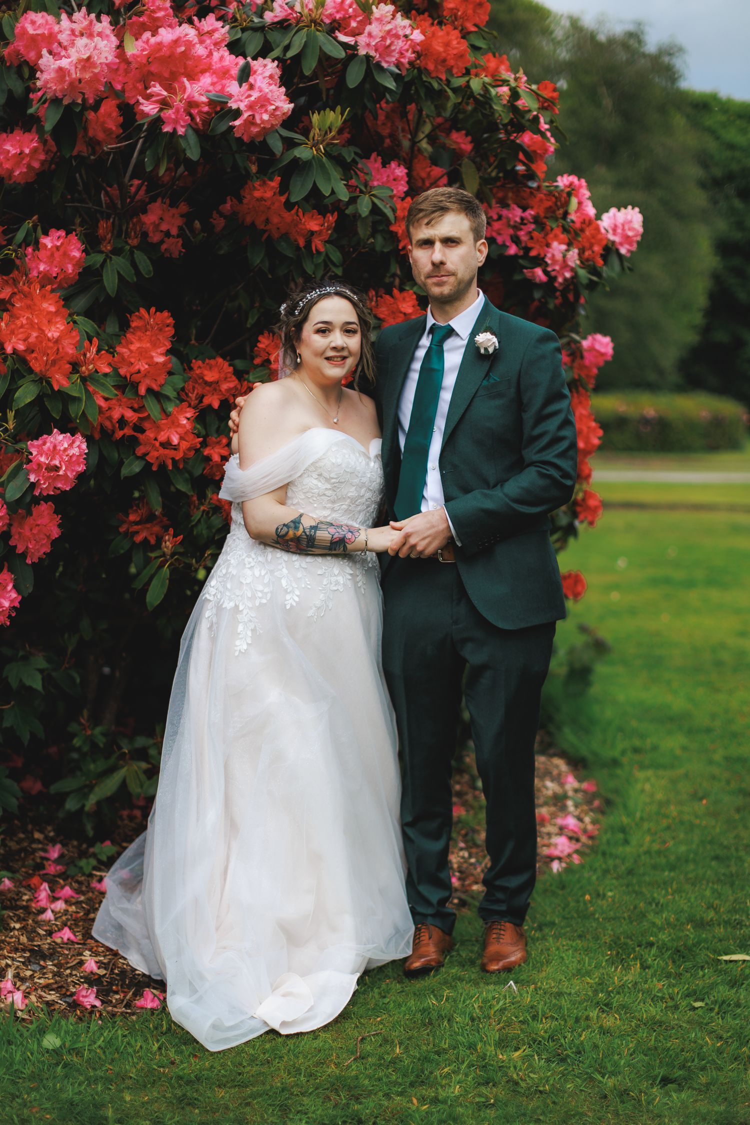 Bride and groom pose before vibrant pink rhododendron blooms, she in white off-shoulder gown, he in dark suit with teal tie.