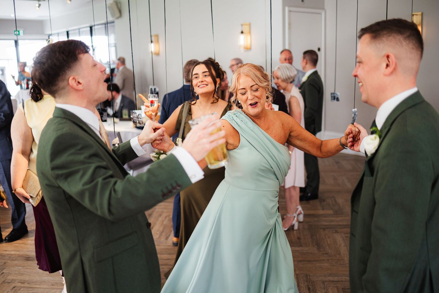 Wedding guests in sage green attire dancing and laughing joyfully at an elegant indoor wedding reception.