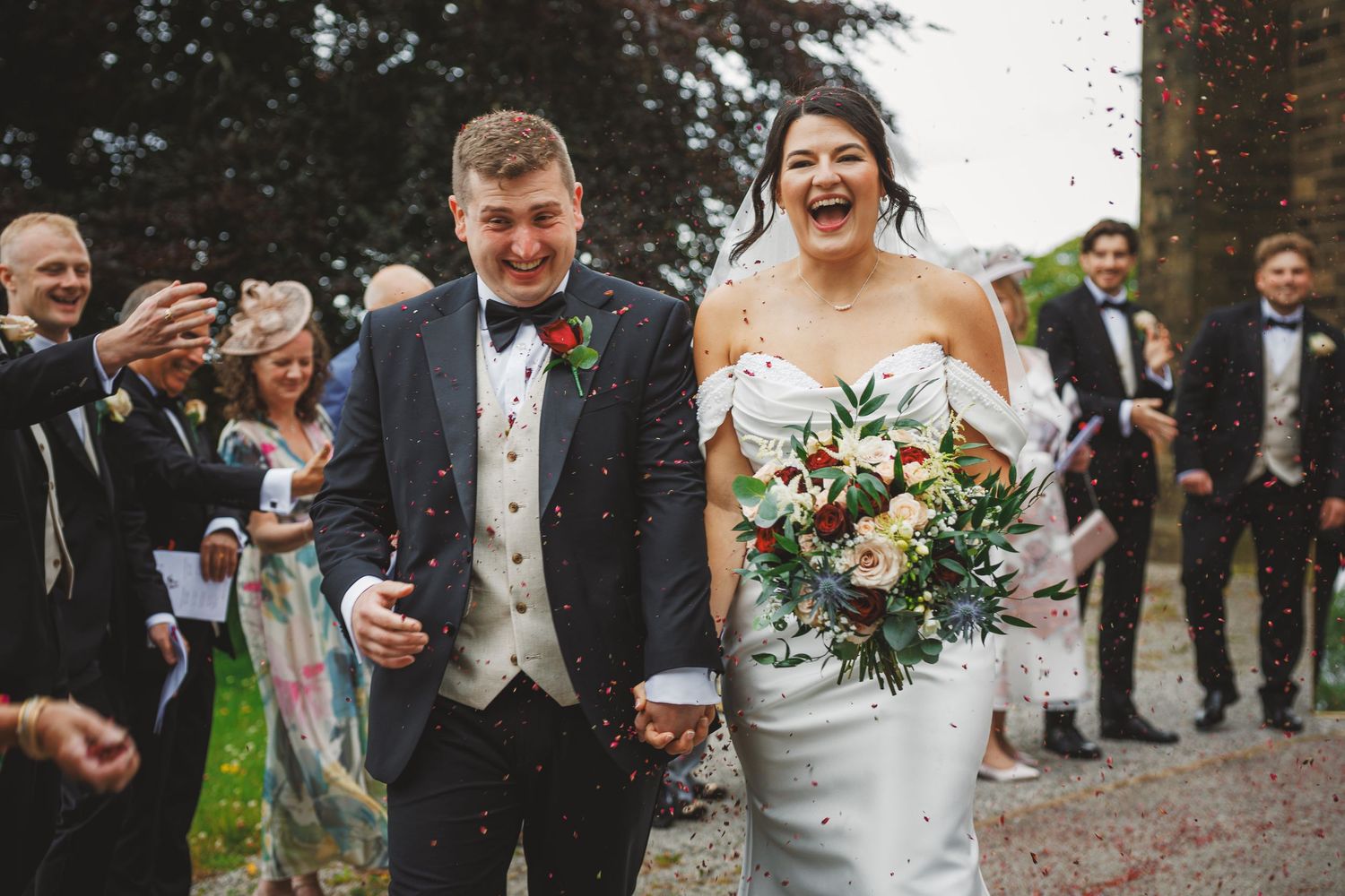 Joyful newlyweds walk through confetti shower, bride holding bouquet in strapless gown, groom in gray suit with red boutonniere.