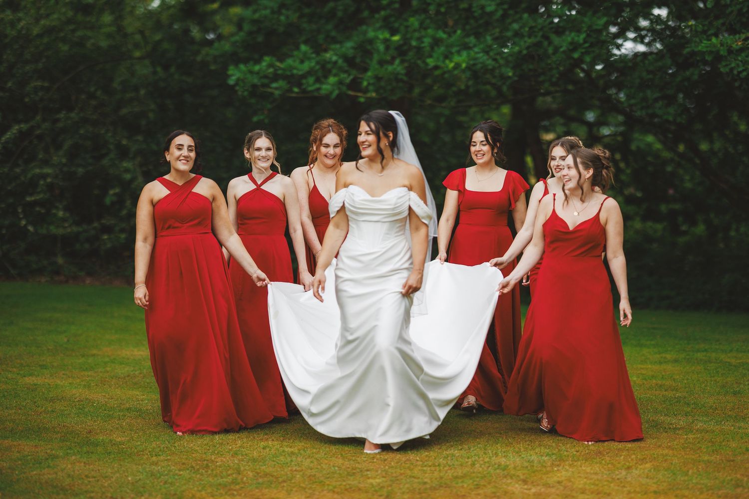 Bride in white gown with five bridesmaids in red dresses walking on green lawn with trees in background.