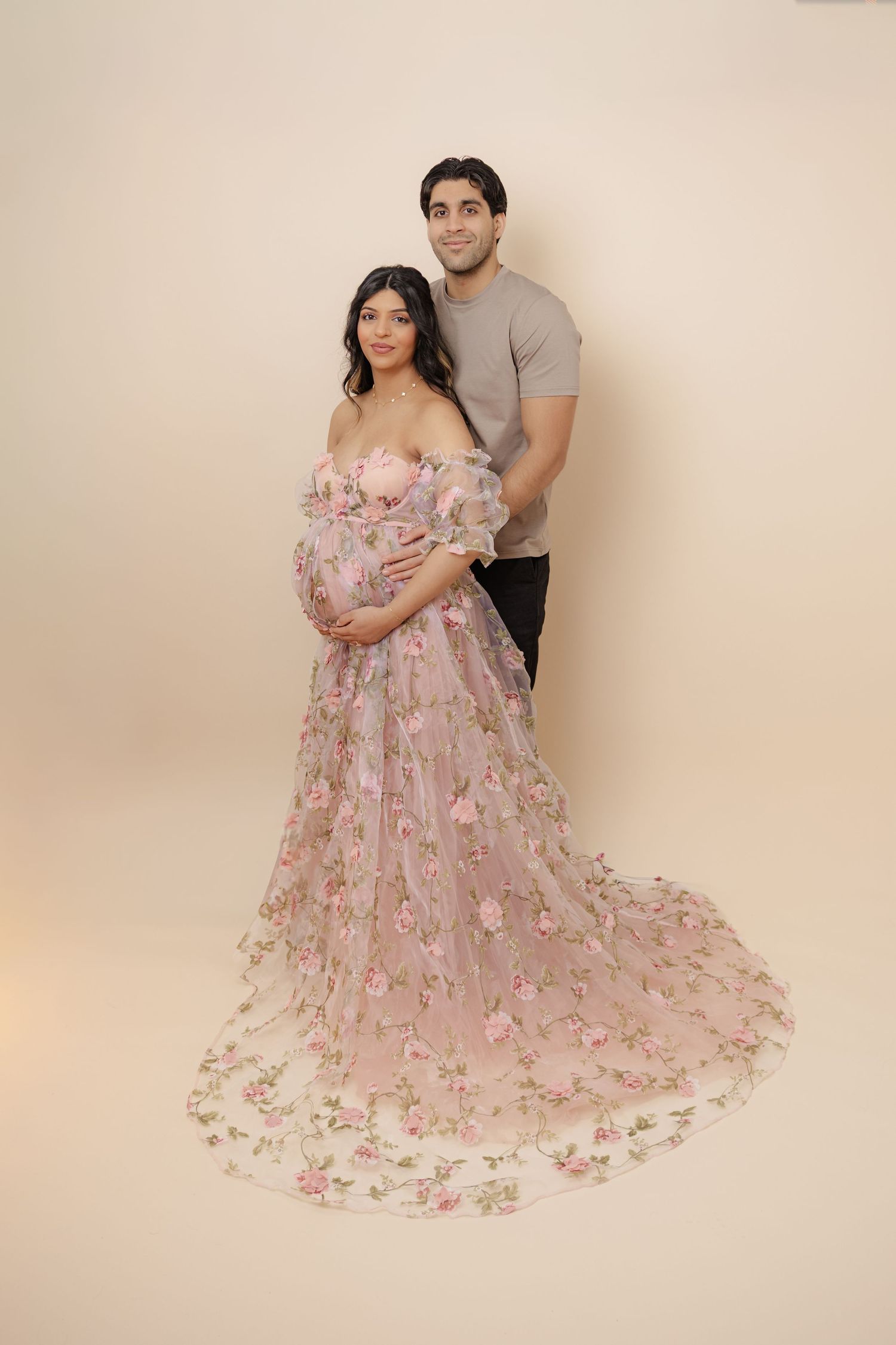 Pregnant woman in floral pink gown with partner during maternity photoshoot on beige background.
