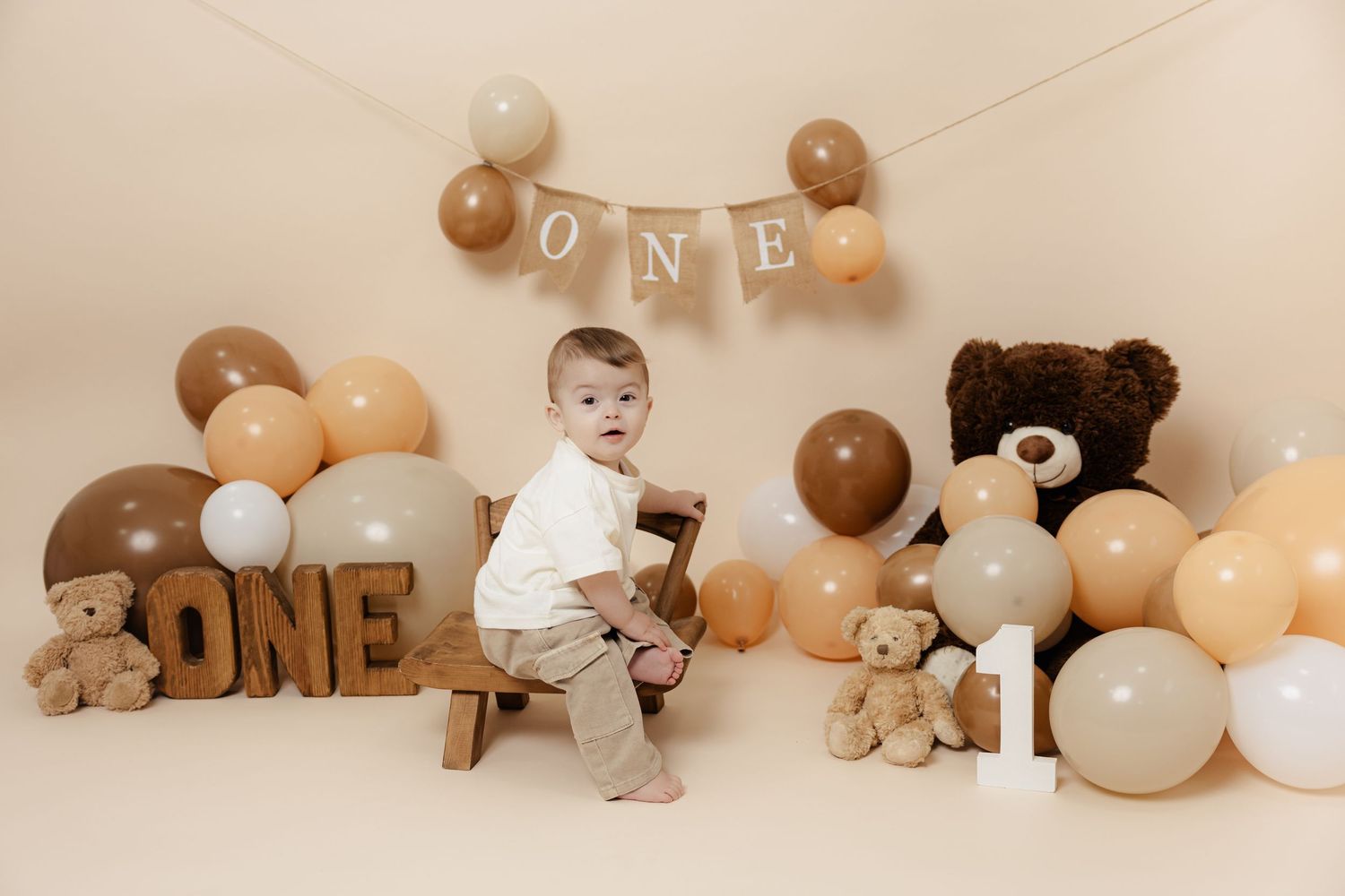 Baby's first birthday cake smash photo with ONE banner, teddy bears, and beige balloons on cream background.