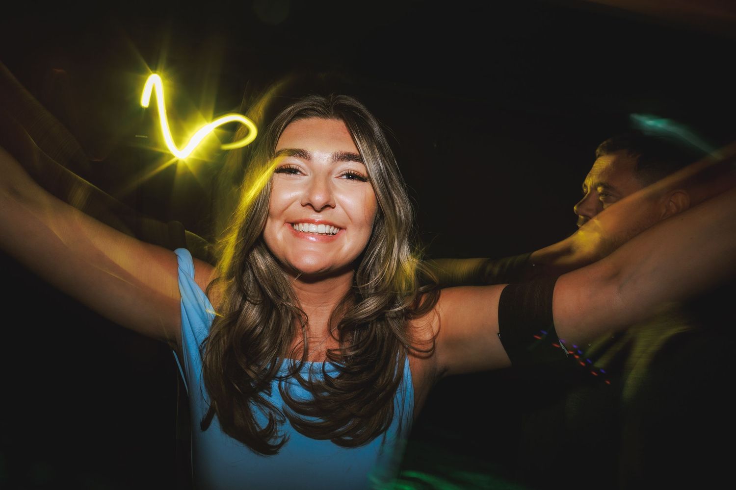 Smiling woman with long brown hair holding glowing light trails in a dark party setting.