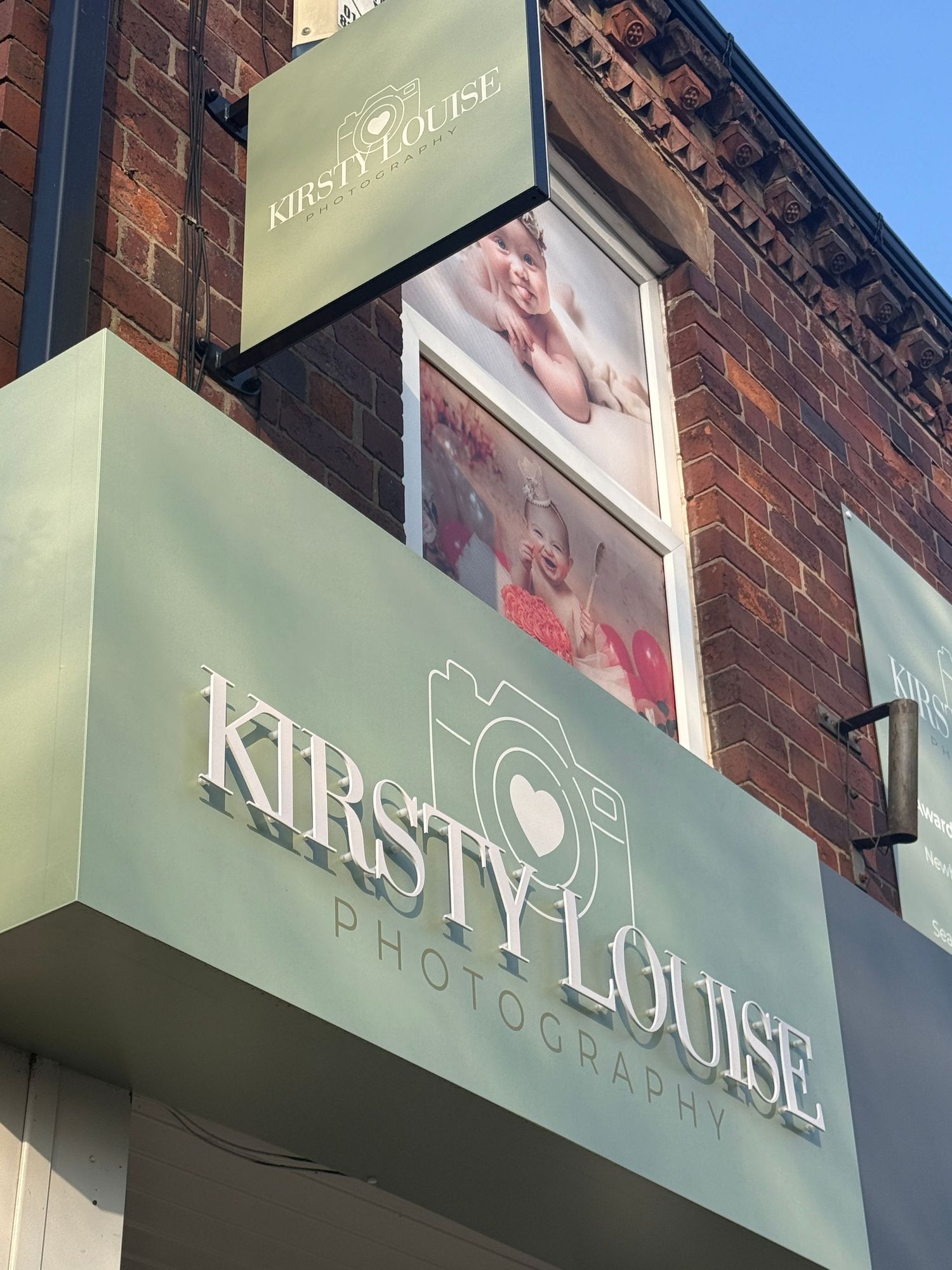 Kirsty Louise Photography studio storefront sign on a brick building with baby portrait displays in the window.