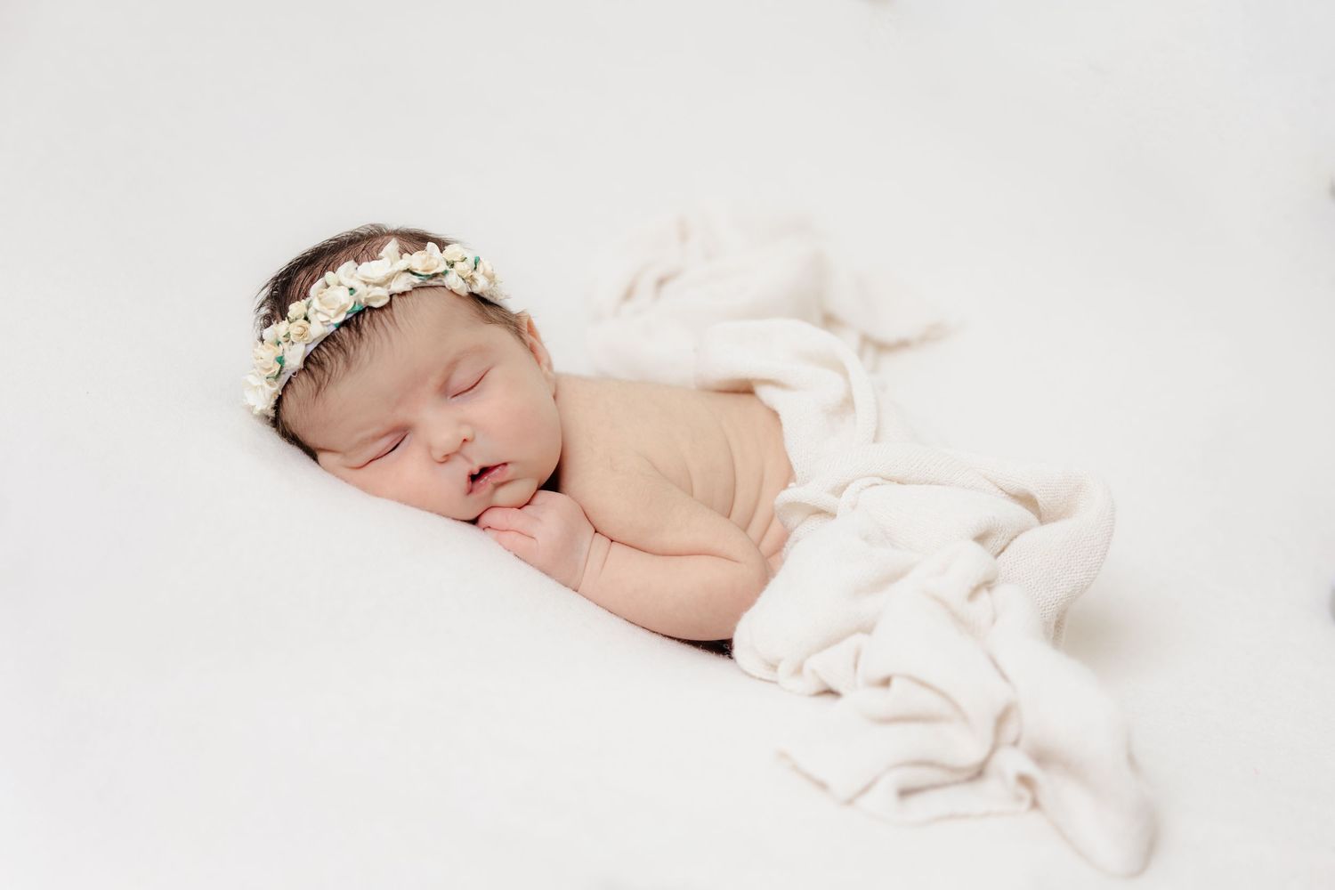 Peaceful newborn baby sleeping on white blanket wearing delicate floral headband in soft studio portrait.