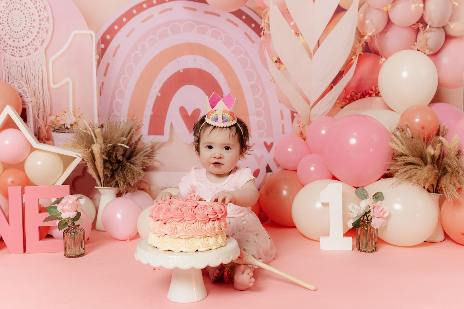 Baby girl in pink tutu sits before a first birthday cake smash setup with balloons, rainbows, and number one decorations.