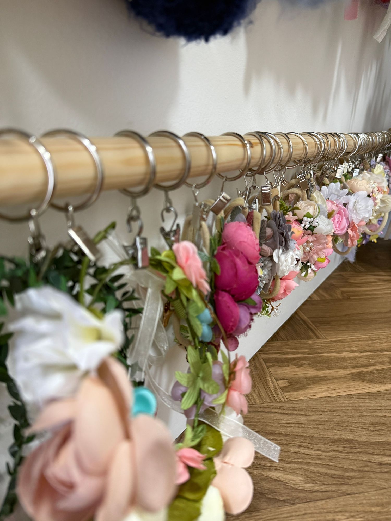 Floral crowns with pink, white, and green flowers hanging on a wooden curtain rod with silver rings.