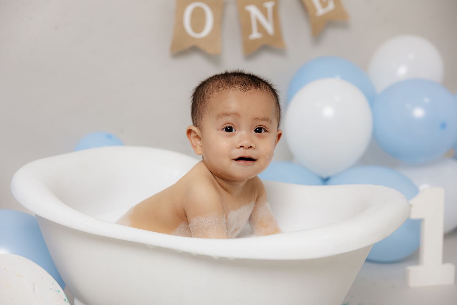 A baby boy sits in a white mini bathtub surrounded by blue and white balloons celebrating his first birthday.