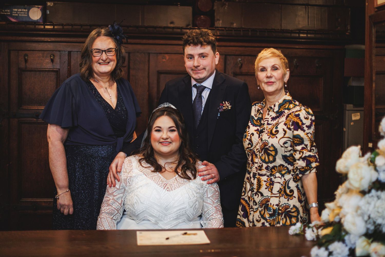 Bride in white lace gown sits at signing table with groom and two female guests at an elegant wedding ceremony.
