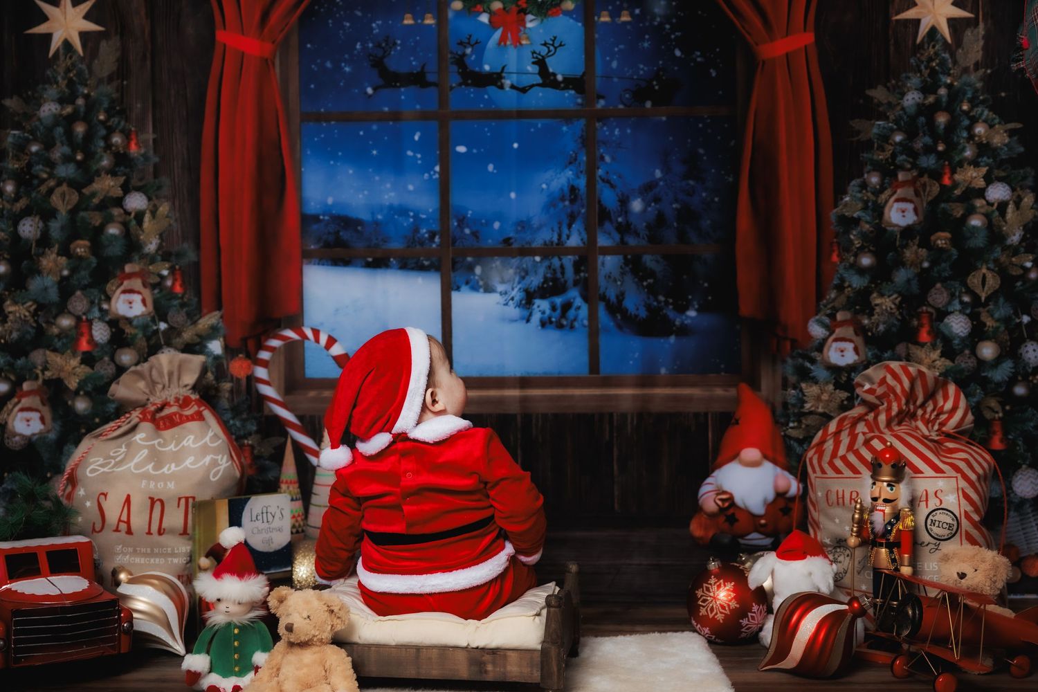Child dressed as Santa sits by a festive window with Christmas trees, toys, and reindeer silhouette in snowy night sky.