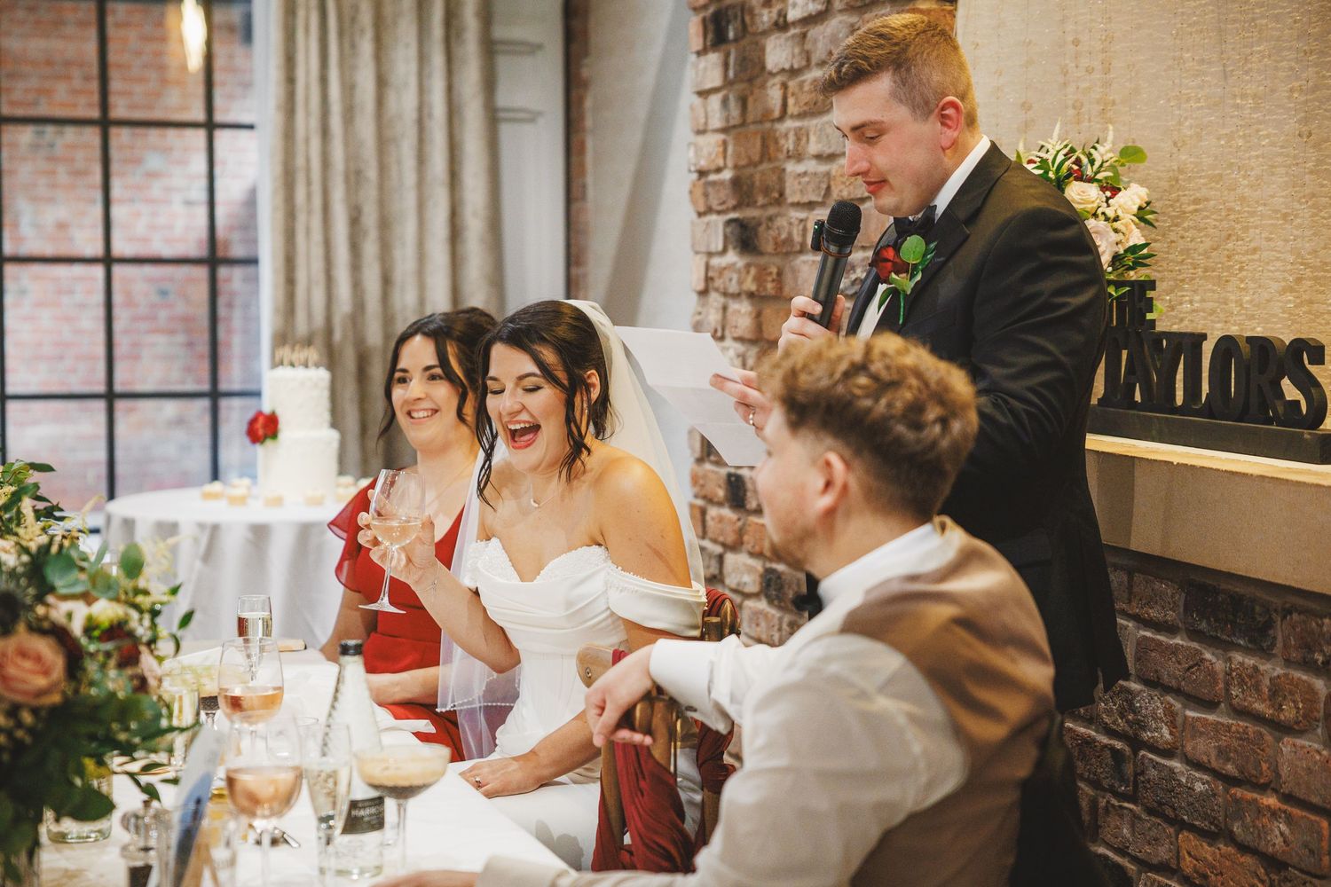 Bride and groom laughing during wedding reception speech at elegant brick-walled venue with floral decorations.