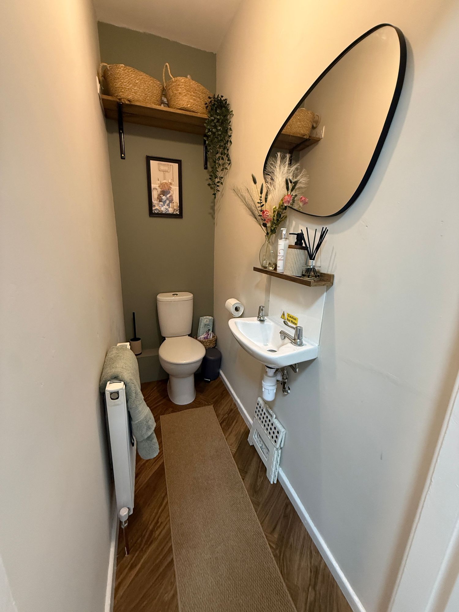 Narrow cloakroom with white toilet, small sink, oval mirror, wooden shelves with baskets, green wall, and wood-effect flooring.