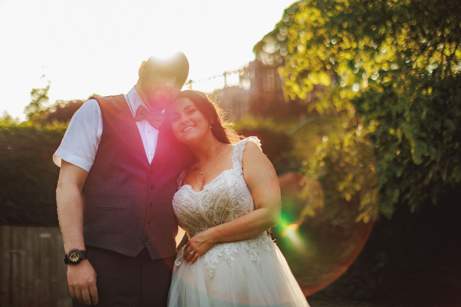 Bride and groom embracing outdoors with golden sunset light and lush green trees in background.