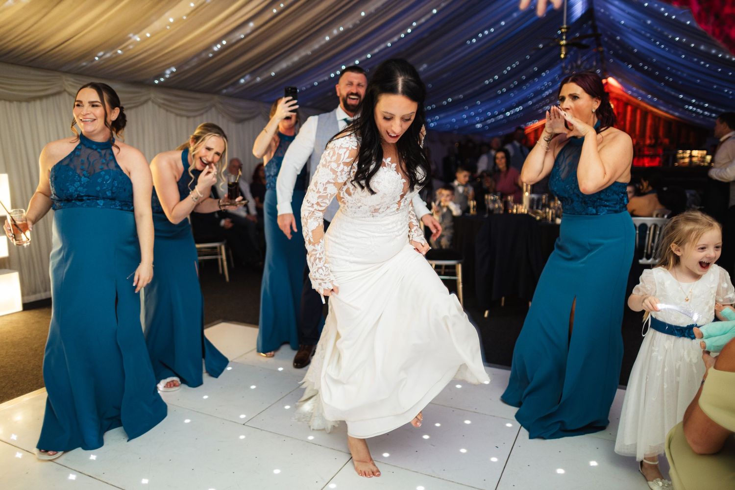 Bride dancing joyfully with bridesmaids in teal dresses on a white dance floor at an elegant wedding reception.