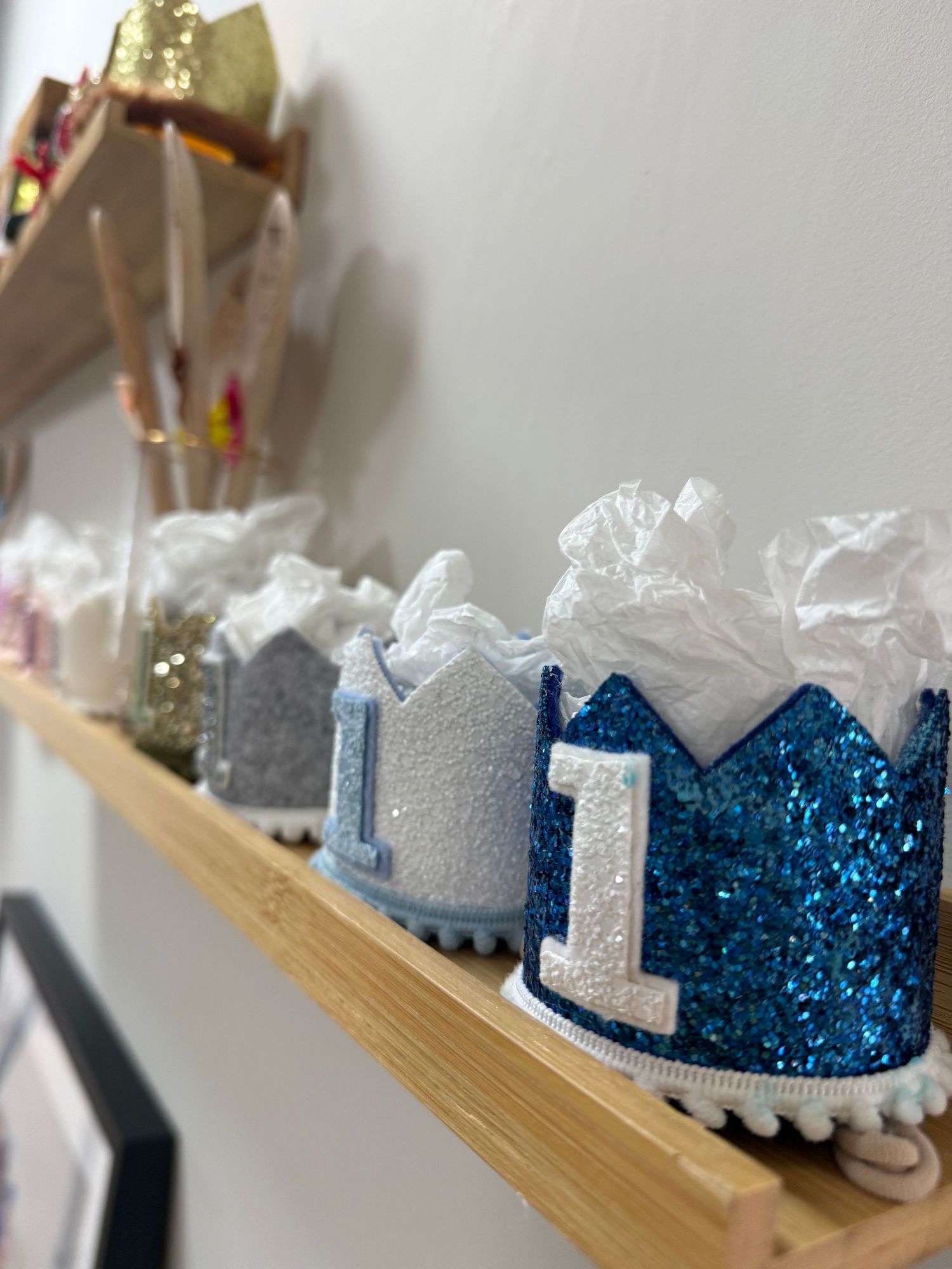 Blue and silver glitter number 1 birthday crowns displayed on a wooden shelf for a first birthday party.