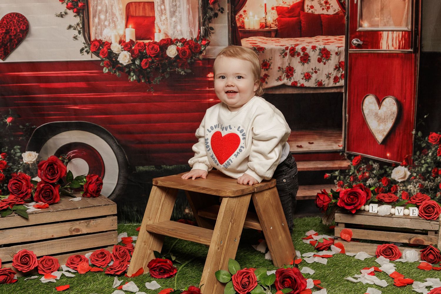 Baby sitting on wooden stool surrounded by red roses and vintage red bus Valentine's Day photo backdrop.