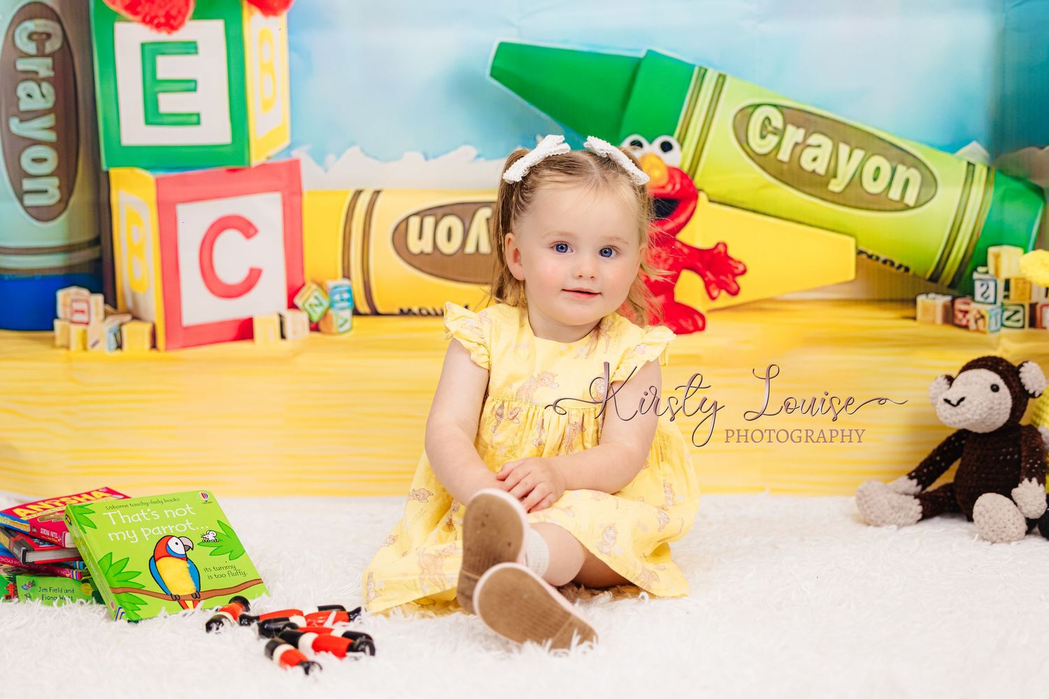 Adorable baby girl in yellow dress sits surrounded by colorful crayon and alphabet block decorations for a photo shoot.