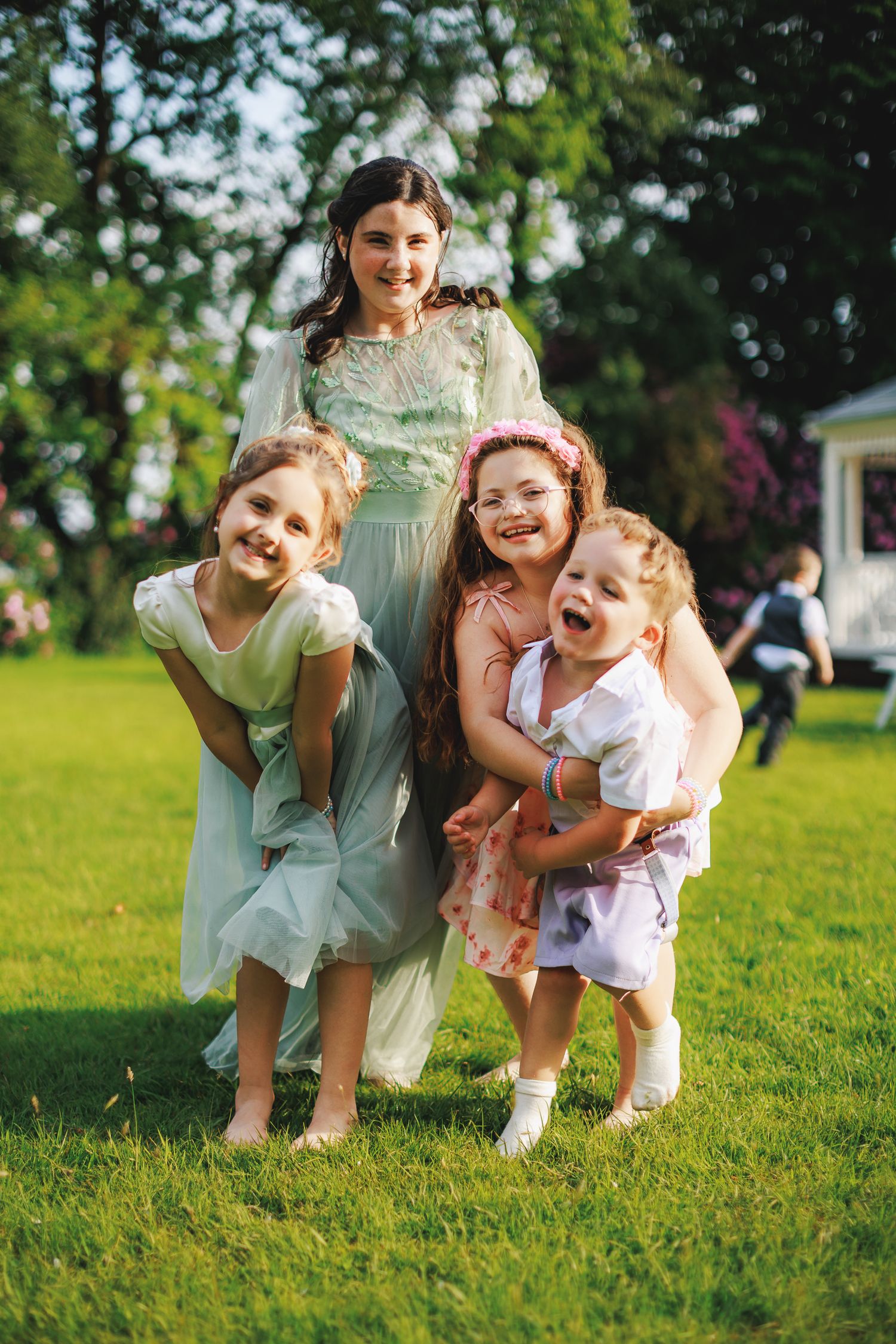 Four joyful girls in pretty dresses laugh and pose together on green grass at an outdoor summer celebration.