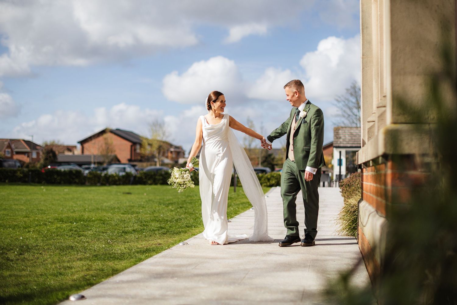 Bride in white jumpsuit and groom in green suit hold hands walking along a path at an outdoor wedding venue.