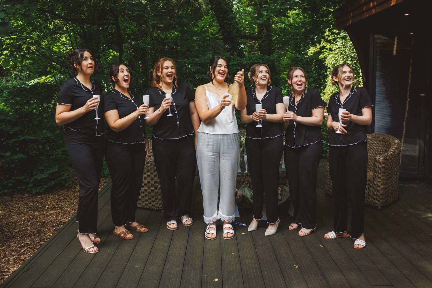 Bride in white pajamas laughing with six bridesmaids in black pajamas holding champagne glasses on a wooden deck.