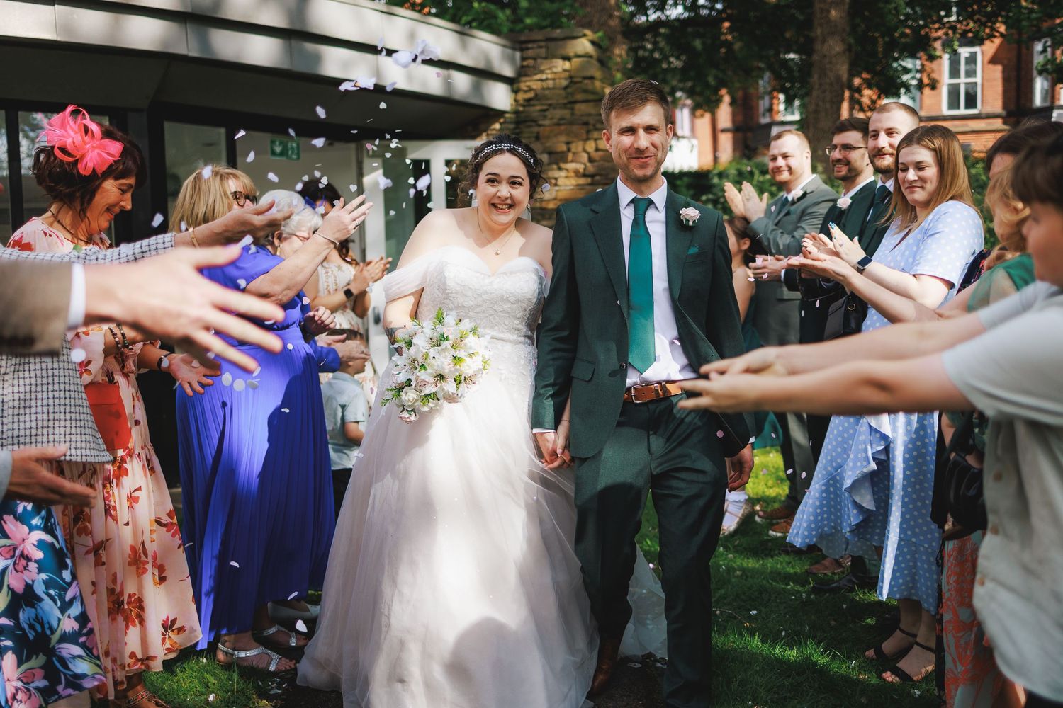 Newlywed couple walks through confetti shower surrounded by smiling wedding guests outdoors near brick building.