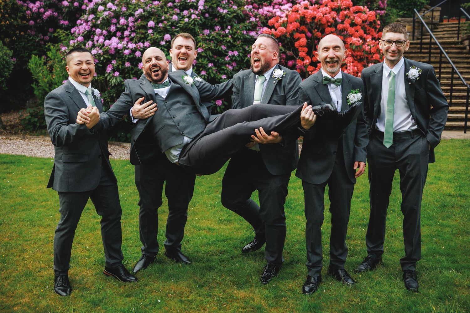 Groomsmen in gray suits laughing while carrying groom horizontally outdoors with colorful flowers in background.