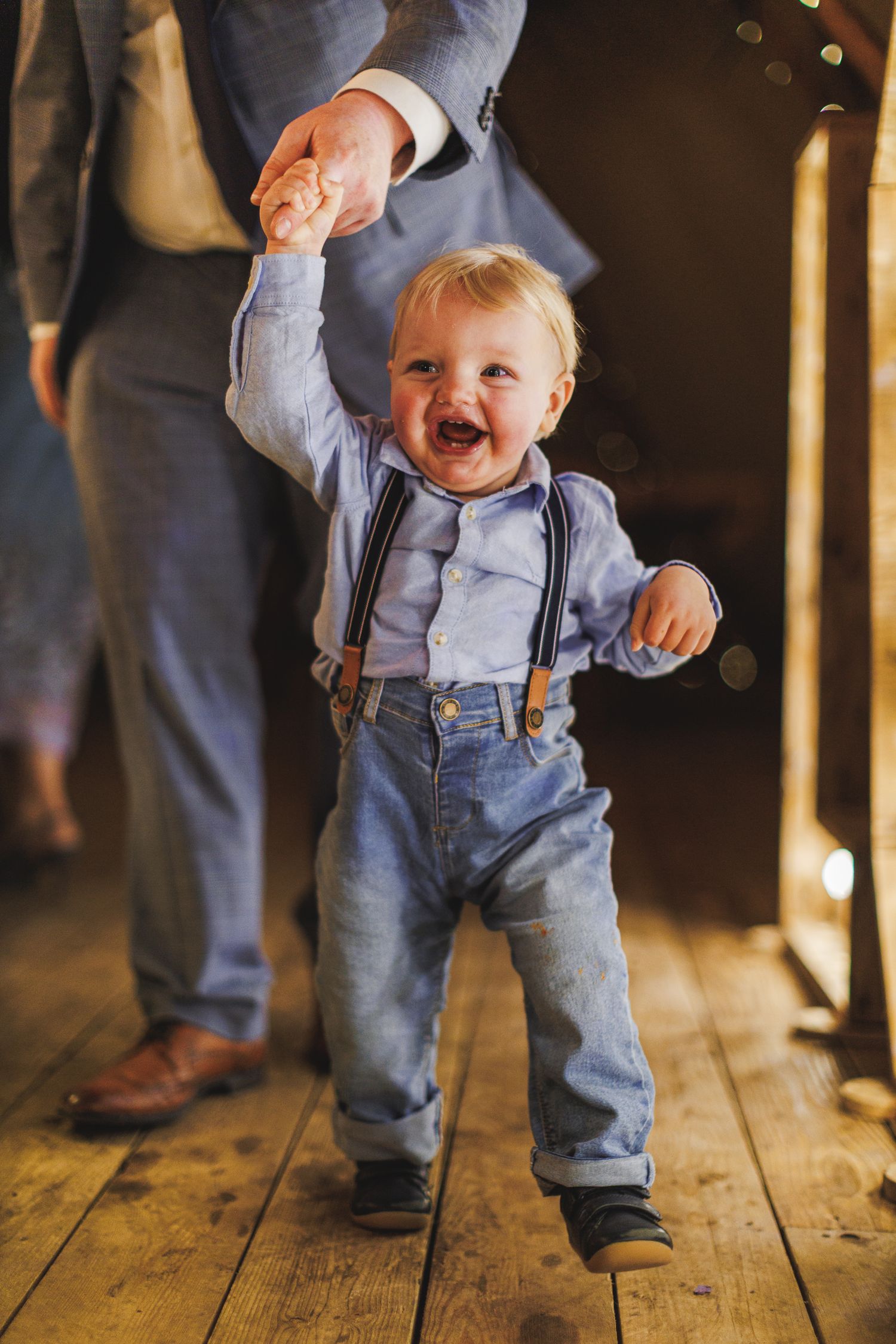 Smiling toddler boy in suspenders and jeans holds an adults hand while standing on a wooden floor.