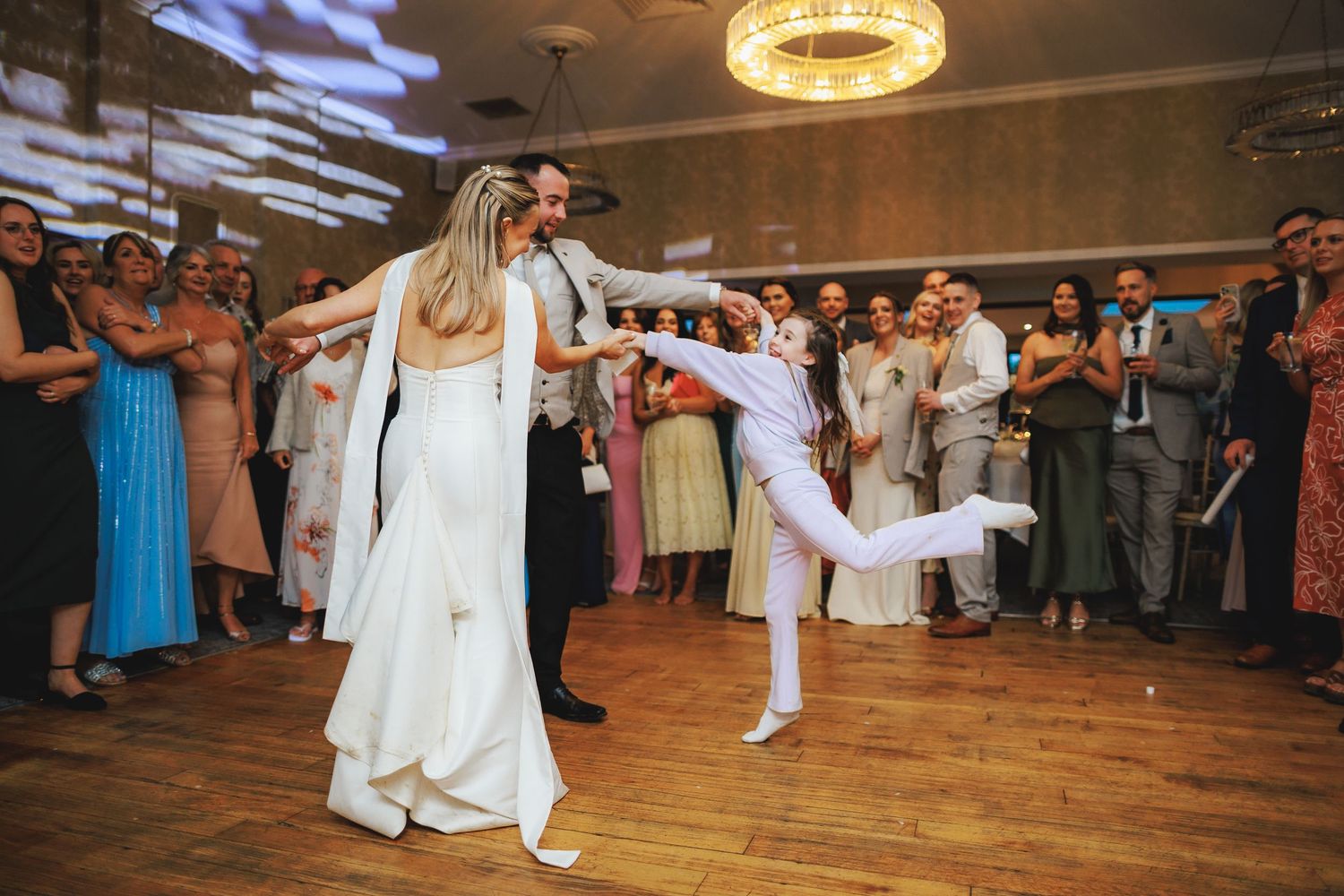 Bride and groom share energetic first dance at wedding reception with guests watching and applauding on wooden dance floor.