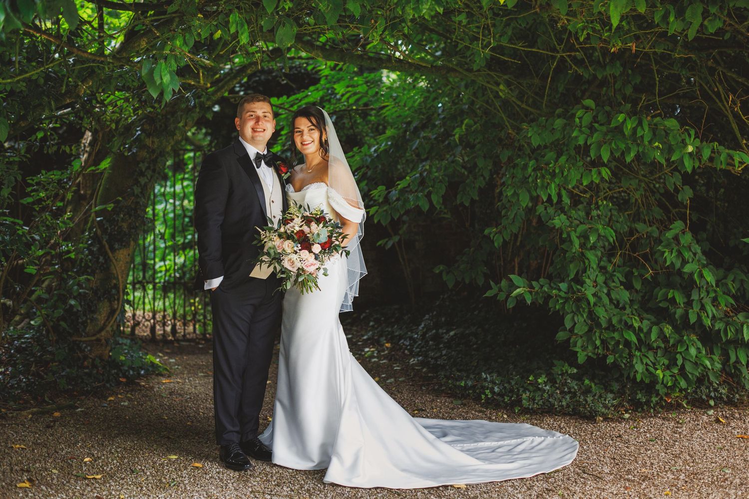 Bride and groom pose under lush green archway, bride in elegant white gown with veil holding floral bouquet.
