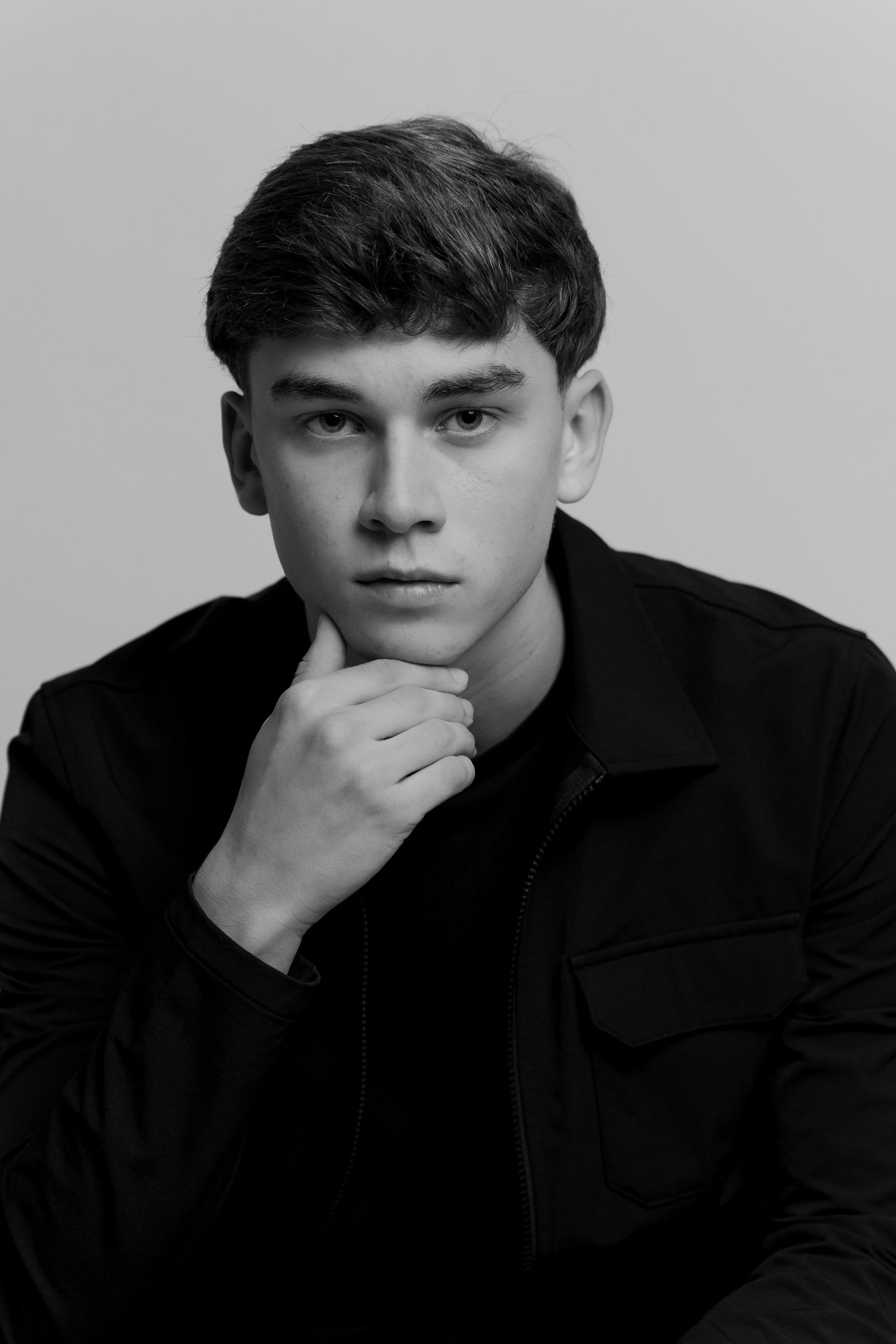 Black and white portrait of a young man in a dark shirt, resting chin on hand, with a thoughtful expression.