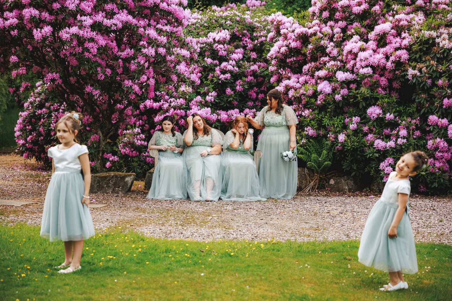 Bridesmaids in sage green gowns pose with bride before pink rhododendron blooms as flower girls play on lawn.