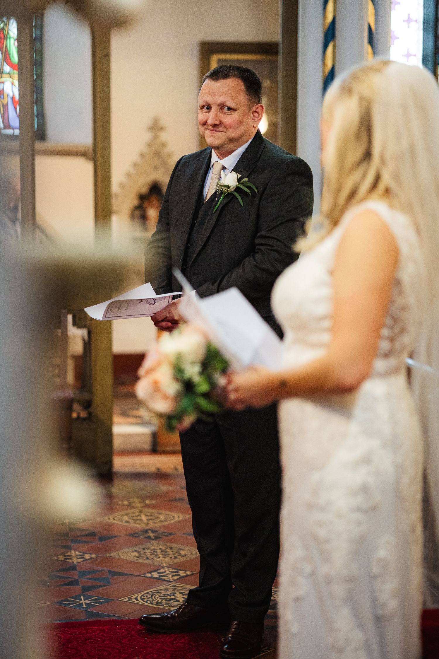 Groom smiling at bride during church wedding ceremony, both holding programs, with stained glass windows in background.