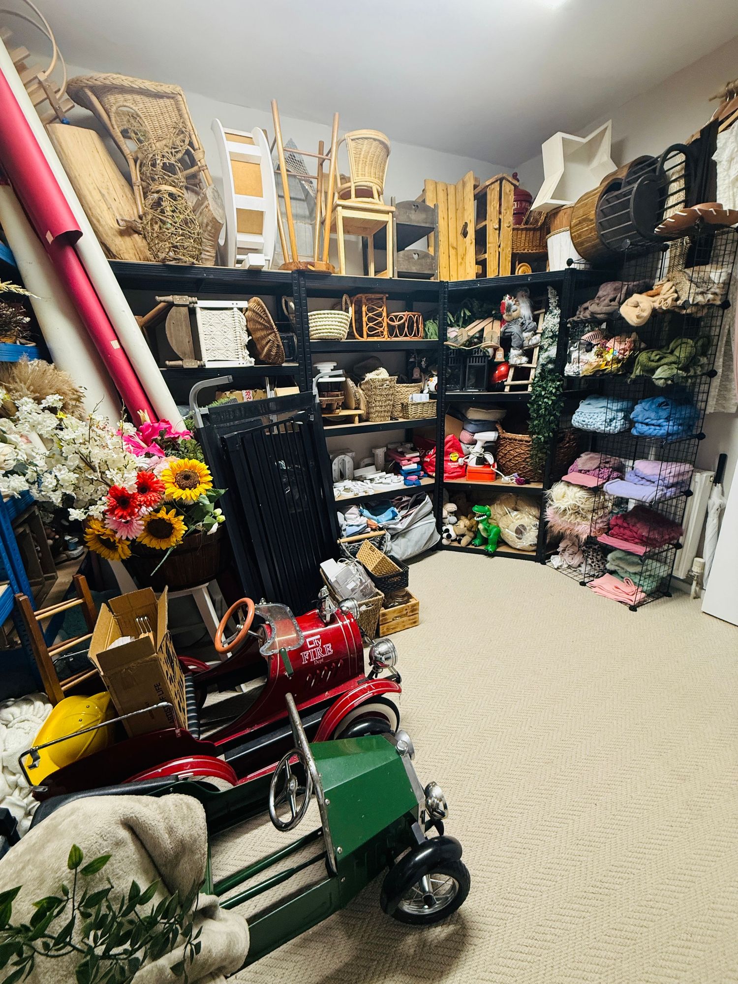 Cluttered storage room with shelves of fabric, baskets, toys, and vintage items including a red pedal car.