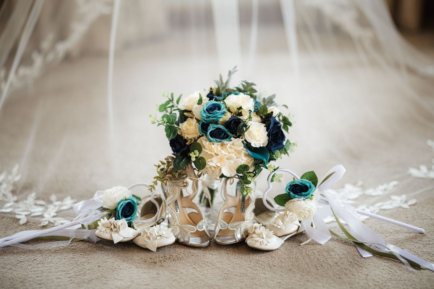 Bridal bouquet with teal and ivory roses beside white heels under a lace veil on a burlap surface.
