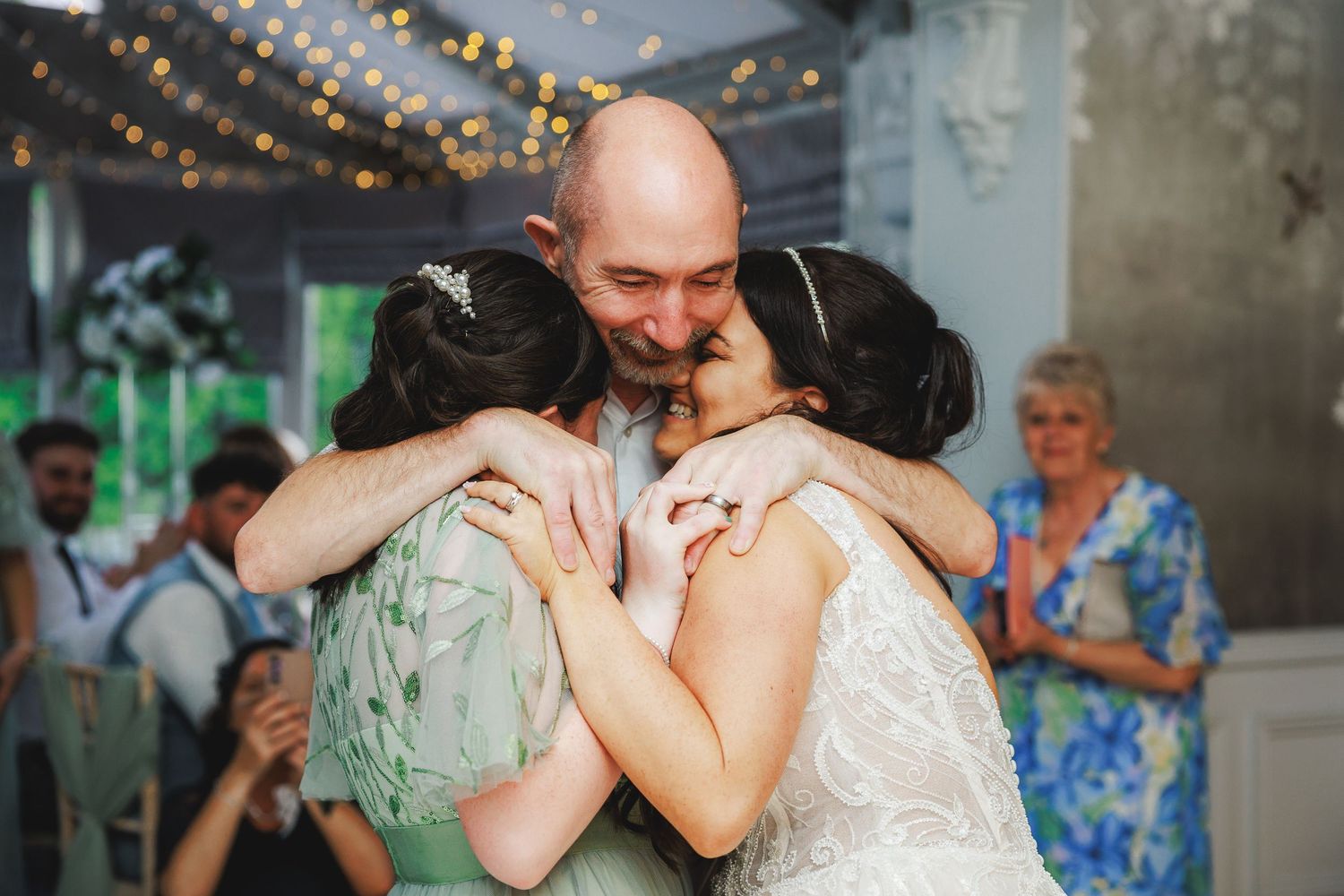 Emotional wedding moment as bald groom hugs bride and bridesmaid in green dress at elegant reception venue.