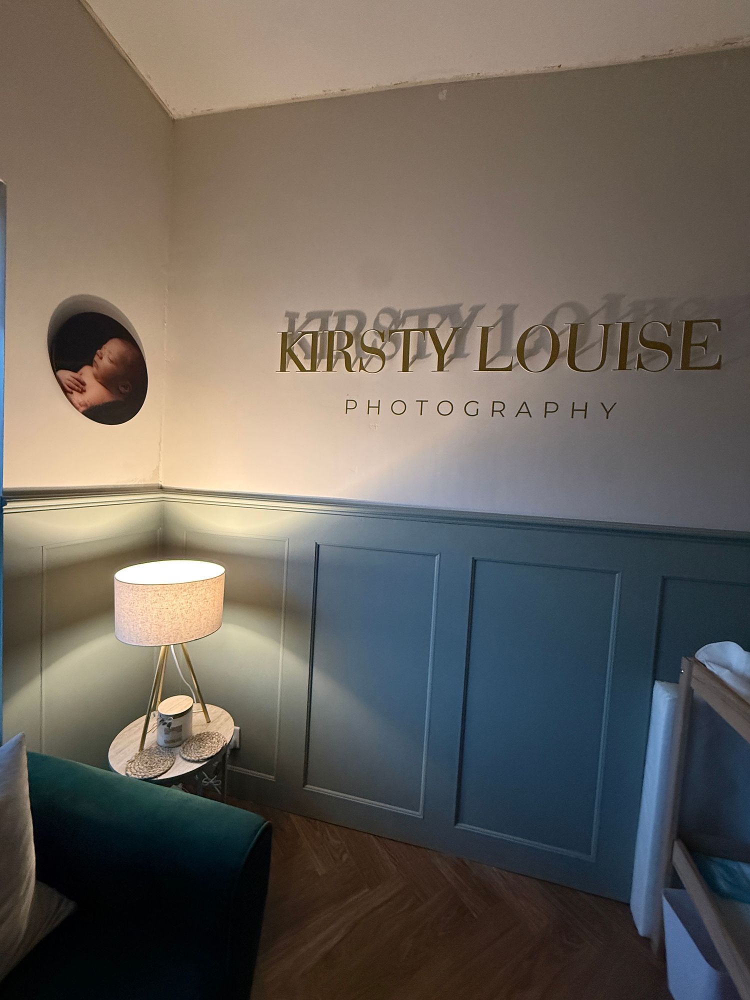 Kirsty Louise Photography studio interior with teal wainscoting, gold lettering, herringbone floor, and a lit table lamp.