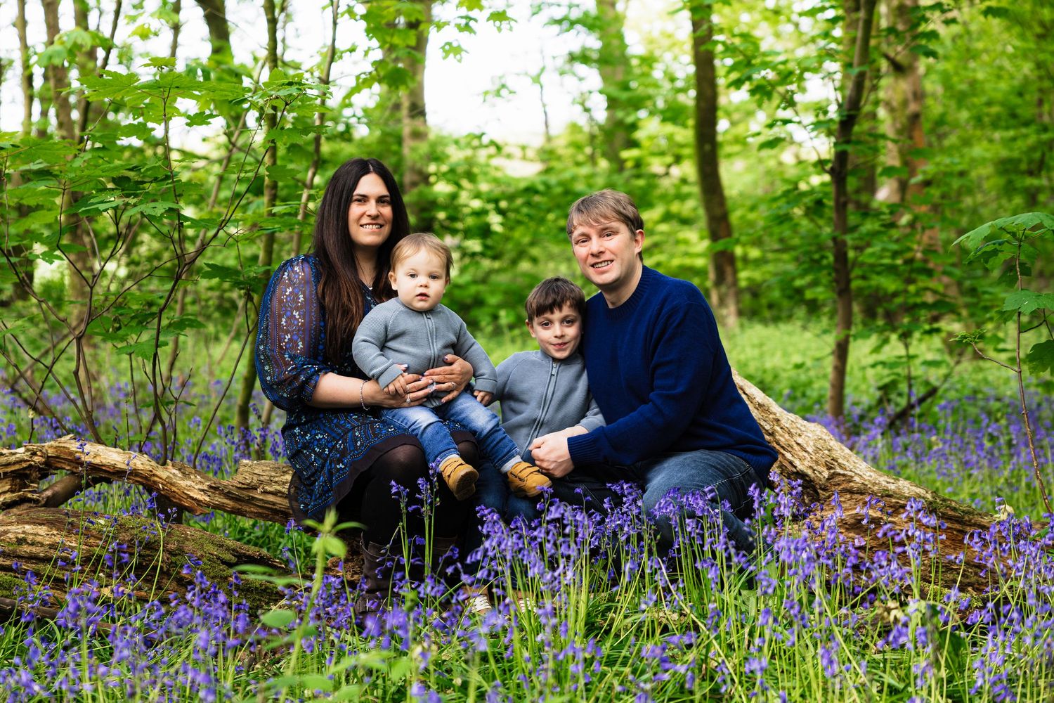 Family of four sitting on a fallen log surrounded by bluebells in a lush green woodland setting.