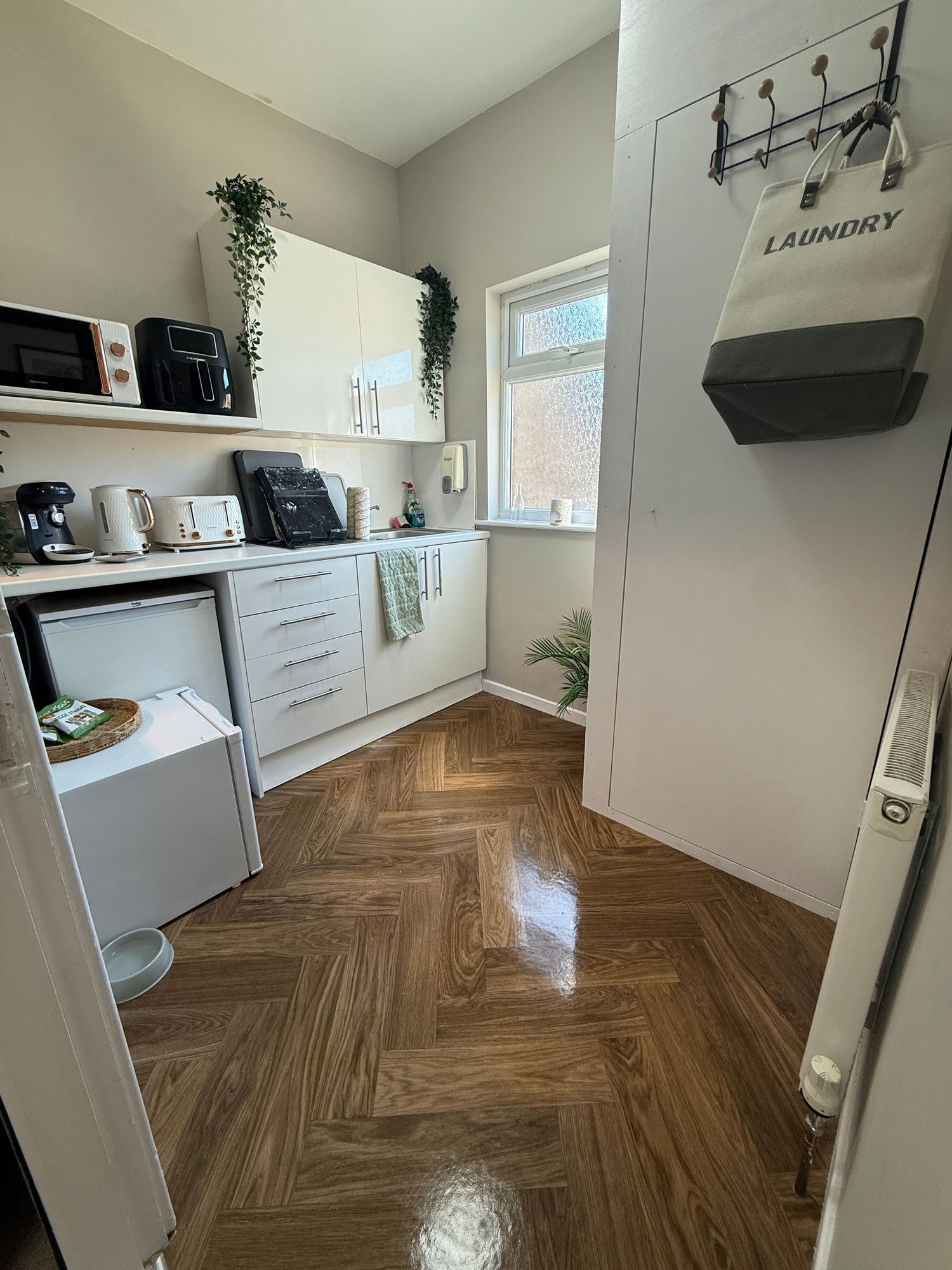 Compact kitchen with herringbone wood floors, white cabinets, open shelving, small appliances, and a laundry sign on the door.