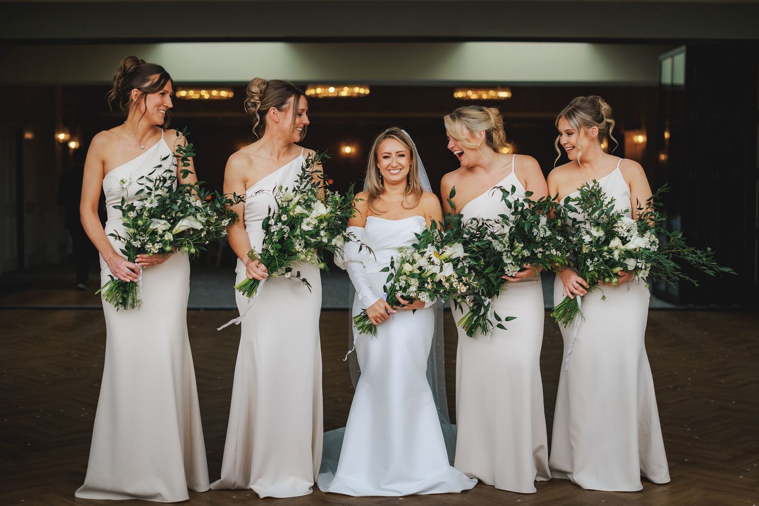 Bride in white gown with four bridesmaids in champagne dresses holding lush greenery bouquets at elegant wedding venue.