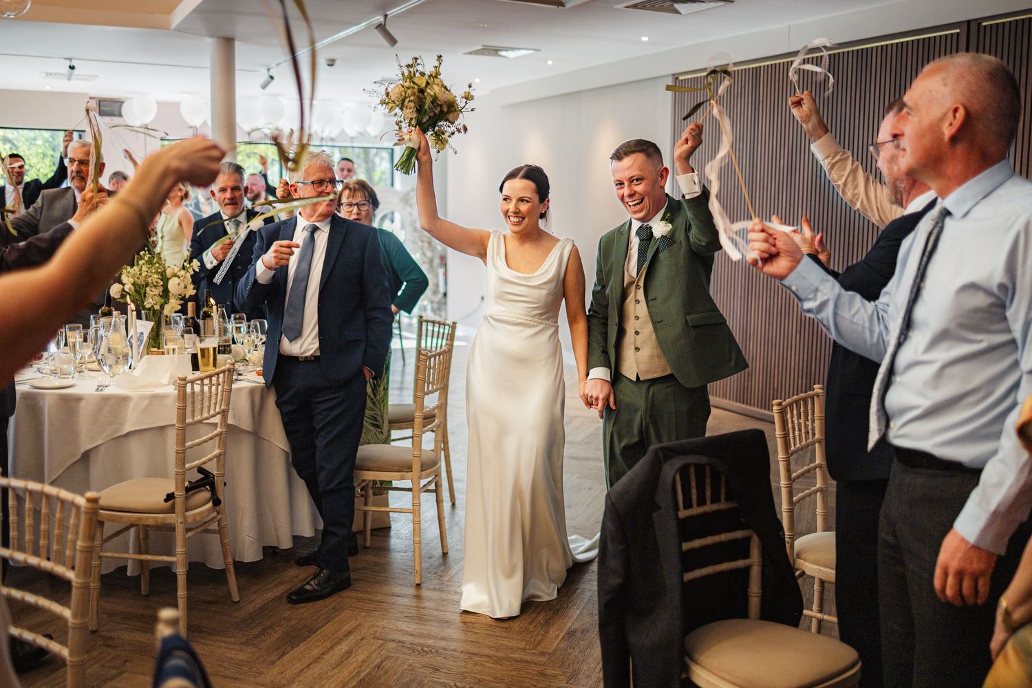 Bride and groom celebrate their wedding reception entrance, guests raising glasses in a bright elegant venue.