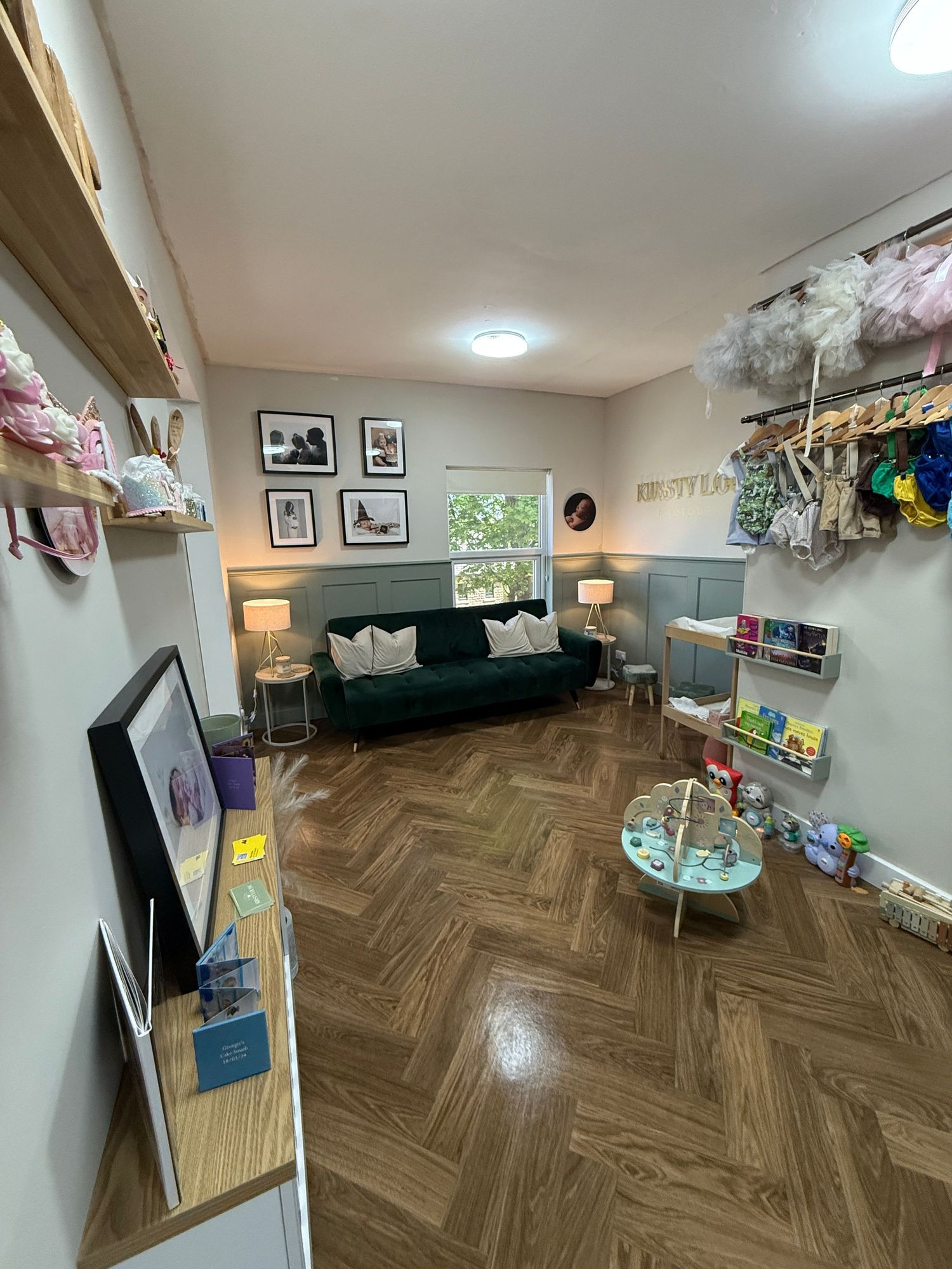Cozy boutique interior with herringbone wood floors, green velvet sofa, wall art, and colorful children's clothing displays.