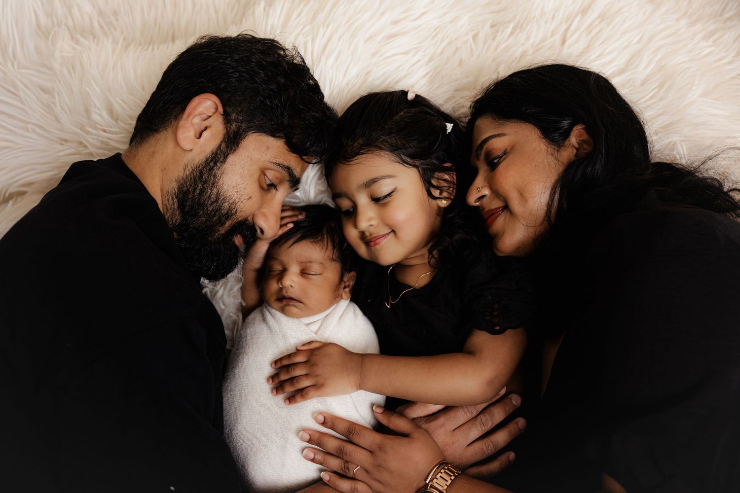 Family of four in matching black outfits cuddling a newborn baby on a white fur backdrop.