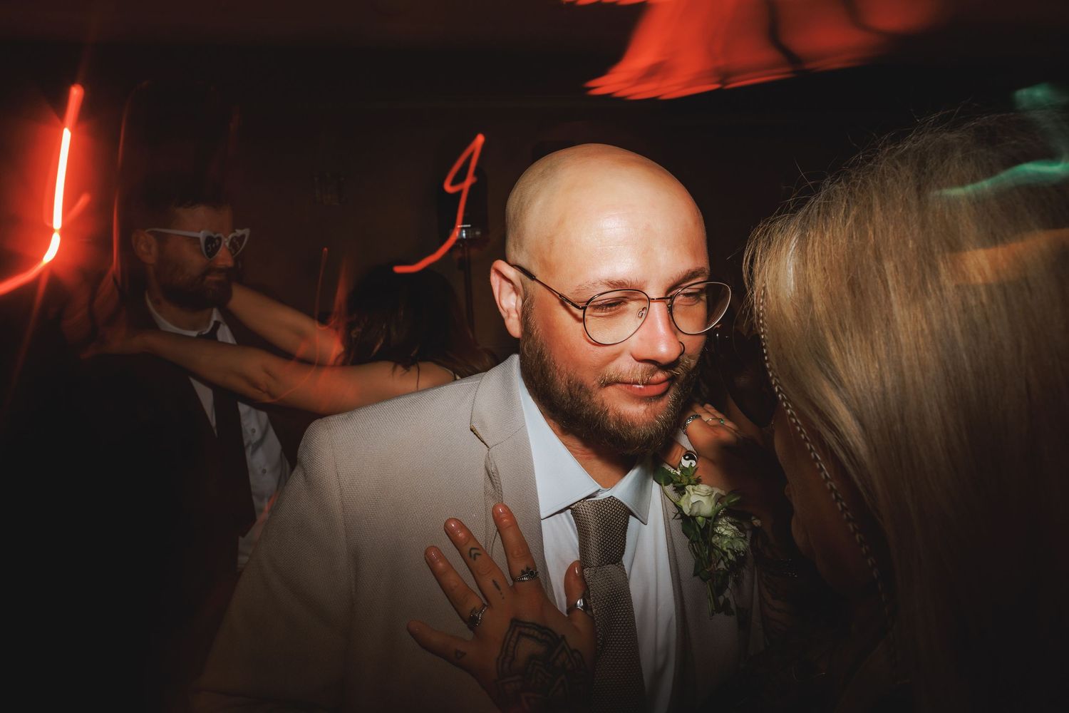 Bald man with glasses in white suit jacket wearing boutonniere at a dimly lit wedding reception dance floor.
